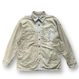 Puede incluir: Chaqueta abotonada de color beige con dos bolsillos en el pecho y un pequeño parche con el logotipo. La chaqueta tiene cuello y mangas largas. La tela parece desgastada, con algo de decoloración. La chaqueta está abotonada y tiene un estilo clásico de ropa de trabajo.