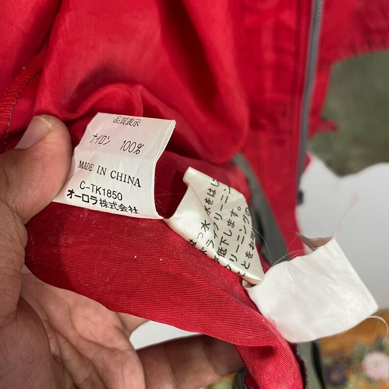Op de afbeelding: Een rood nylon jack met een wit label waarop staat: "Kwaliteitsweergave", "Nylon 100%", "Gemaakt in China", "C-TK1850" en "Aurora Co., Ltd."