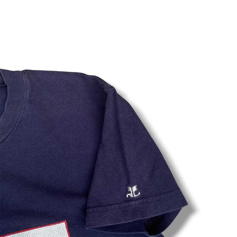 K&ouml;nnte beinhalten: Ein dunkelblaues T-Shirt mit kurzen &Auml;rmeln und einem kleinen, wei&szlig; gestickten Logo auf dem &Auml;rmel. Das Logo besteht aus zwei stilisierten, gespiegelten Buchstaben, die den Zeichen AC &auml;hneln. Der Stoff hat eine feine, sichtbare Textur.