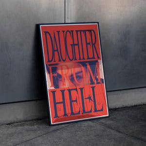 Op de afbeelding: Een rechthoekige ingelijste poster met een rode achtergrond en de donkerblauwe tekst DAUGHTER FROM HELL. Het ontwerp toont een gedeeltelijk gezicht met intense ogen achter de tekst en een klein sterretje verwerkt in het woord HELL.