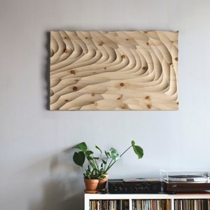 Puede incluir: Una pieza de arte de pared de madera con un diseño tallado en capas. La madera es de color marrón claro y el diseño recuerda a olas o ondas.