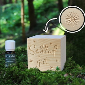 Puede incluir: Un difusor de madera en forma de cubo con el texto "Schlaf gut" y estrellas talladas en la superficie. También se incluye una pequeña botella de aceite esencial.