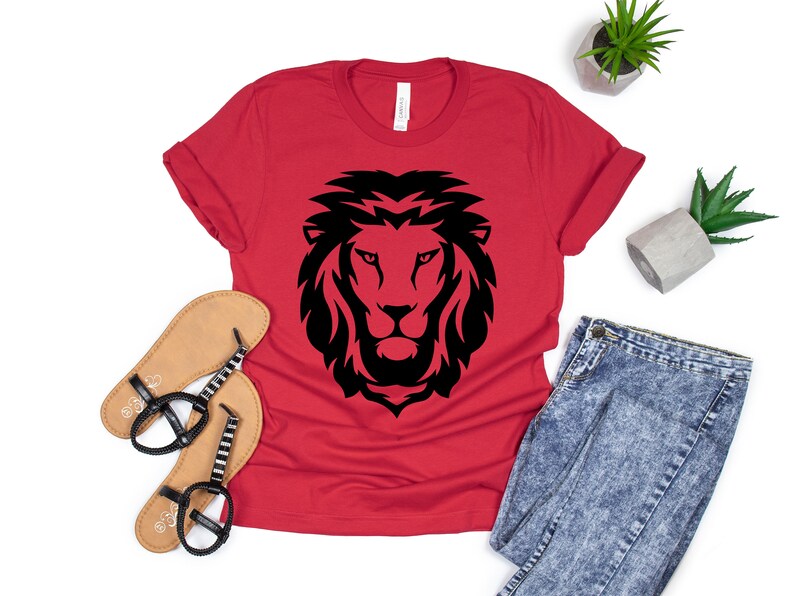 K&ouml;nnte beinhalten: Ein rotes T-Shirt mit einem schwarzen L&ouml;wenkopf-Design. Das Shirt wird mit Sandalen, einer Jeans und kleinen Topfpflanzen pr&auml;sentiert. Die L&ouml;wen-Grafik ist auf dem Shirt zentriert, mit einer detaillierten, stilisierten M&auml;hne und Gesichtsz&uuml;gen.