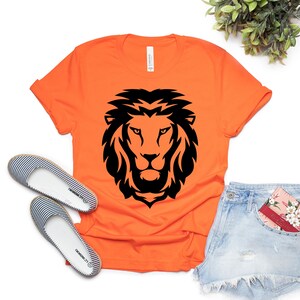 K&ouml;nnte beinhalten: Orangefarbenes T-Shirt mit einem schwarzen L&ouml;wenkopf-Design. Das Shirt wird mit Jeansshorts und gestreiften Ballerinas kombiniert. Eine florale Karte befindet sich in der Hosentasche. Das Shirt ist aus weichem Material.