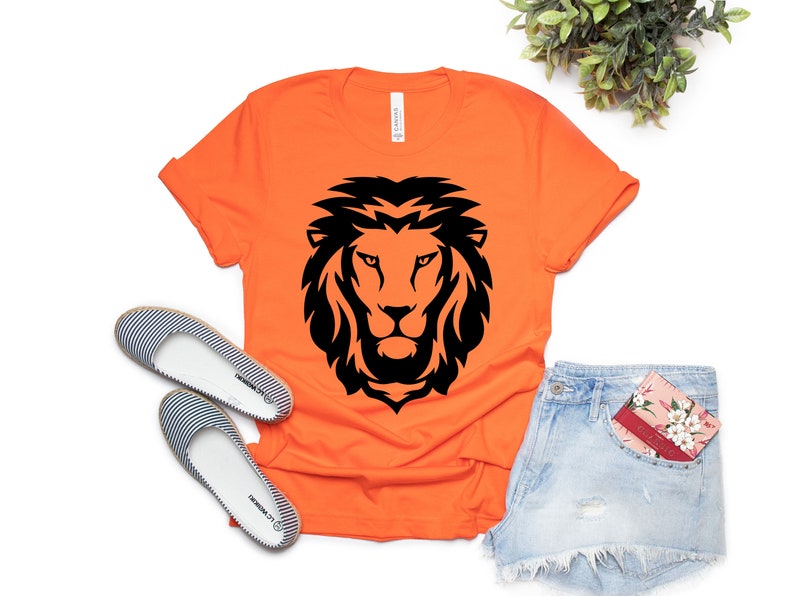 K&ouml;nnte beinhalten: Orangefarbenes T-Shirt mit einem schwarzen L&ouml;wenkopf-Design. Das Shirt wird mit Jeansshorts und gestreiften Ballerinas kombiniert. Eine florale Karte befindet sich in der Hosentasche. Das Shirt ist aus weichem Material.