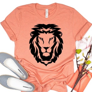 K&ouml;nnte beinhalten: Pfirsichfarbenes T-Shirt mit einem auff&auml;lligen schwarzen L&ouml;wenkopf-Motiv. Das Shirt ist mit hochgekrempelten &Auml;rmeln versehen und wird mit gestreiften Ballerinas und einer korallenfarbenen Clutch kombiniert. Das Design, das sich auf dem Shirt befindet, zeigt die detaillierte M&auml;hne und die Gesichtsz&uuml;ge des L&ouml;wen.