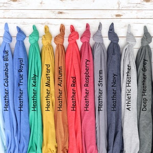 K&ouml;nnte beinhalten: Eine Sammlung gefalteter T-Shirts in verschiedenen Farben, darunter Heather Ice Blue, Heather Columbia Blue, Heather True Royal, Heather Kelly, Heather Mustard, Heather Autumn, Heather Red, Heather Raspberry, Heather Storm, Heather Navy, Athletic Heather, Deep Heather Grey und Dark Grey Heather. Jedes Shirt hat ein Etikett mit seinem Farbnamen.