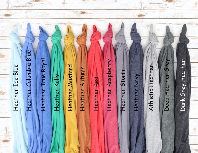 K&ouml;nnte beinhalten: Eine Sammlung gefalteter T-Shirts in verschiedenen Farben, darunter Heather Ice Blue, Heather Columbia Blue, Heather True Royal, Heather Kelly, Heather Mustard, Heather Autumn, Heather Red, Heather Raspberry, Heather Storm, Heather Navy, Athletic Heather, Deep Heather Grey und Dark Grey Heather. Jedes Shirt hat ein Etikett mit seinem Farbnamen.