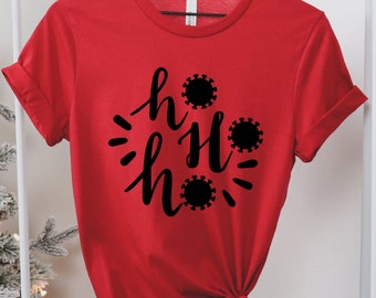 May include: A red t-shirt with the text "ho ho ho" in a black script font, with each "o" replaced by a black coronavirus graphic. The shirt is hanging on a white rack.