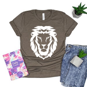 K&ouml;nnte beinhalten: Braunes T-Shirt mit wei&szlig;em L&ouml;wenkopf-Motiv. Das Bild zeigt einen floralen Planer und Jeans. Eine kleine Topfpflanze ist ebenfalls zu sehen.