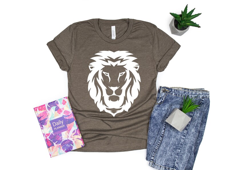 K&ouml;nnte beinhalten: Braunes T-Shirt mit wei&szlig;em L&ouml;wenkopf-Motiv. Das Bild zeigt einen floralen Planer und Jeans. Eine kleine Topfpflanze ist ebenfalls zu sehen.