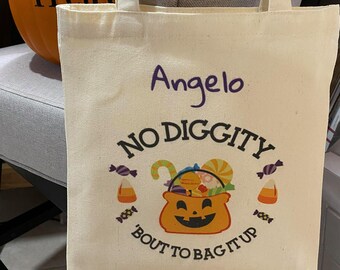 Può includere: Una borsa in tela color crema con il nome Angelo in viola sopra la scritta nera NO DIGGITY 'BOUT TO BAG IT UP. Il design presenta l'immagine di un secchiello a forma di zucca riempito con caramelle colorate.