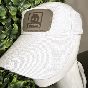 Puede incluir: Visera blanca con un parche marrón con un gráfico de una casa y la palabra "SOLD". La visera está hecha de una tela texturizada y tiene una visera curva. El parche también incluye el texto "Best Sellers to".