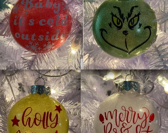 Può includere: Quattro palline di Natale con glitter e diversi design. Una è rossa con testo bianco che dice "Baby it's cold outside". Una è verde con un viso di Grinch nero. Una è gialla con testo rosso che dice "holly jolly". Una è bianca con testo rosso che dice "merry & bright".