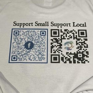 Könnte beinhalten: Weißes T-Shirt mit dem schwarzen Text "Support Small Support Local". Unter dem Text sind zwei QR-Codes aufgedruckt. Der linke QR-Code hat einen blauen Rahmen und ein Facebook-Logo. Der rechte QR-Code hat ein farbenfrohes Design.
