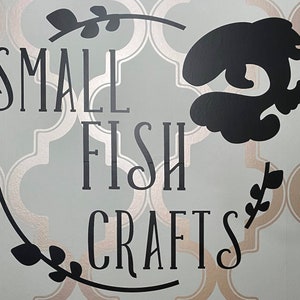 Pu&ograve; includere: Un cartello in bianco e nero con il testo "SMALL FISH CRAFTS" in un carattere decorativo. Il cartello &egrave; su uno sfondo blu chiaro con un motivo dorato.