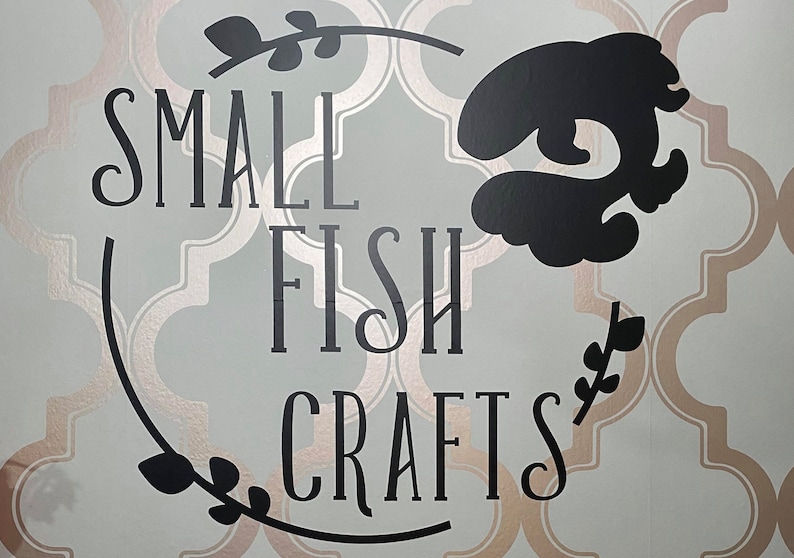 Pu&ograve; includere: Un cartello in bianco e nero con il testo "SMALL FISH CRAFTS" in un carattere decorativo. Il cartello &egrave; su uno sfondo blu chiaro con un motivo dorato.