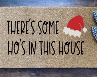 May include: A tan doormat with the text "THERE'S SOME HO'S IN THIS HOUSE" in black. A red Santa hat graphic is on the right. A pair of black and white patterned shoes with red accents are on the right.