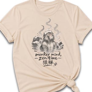 May include: A soft cream-colored t-shirt featuring a graphic of a monkey relaxing in a hot spring with a towel on its head. The text reads monkey mind, zen time, and includes Japanese characters and a small paw print graphic below the monkey.