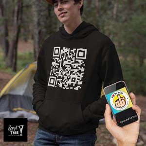 May include: A black hoodie with a white QR code printed on the front. The QR code is a square with a black and white pattern. The hoodie is being worn by a person in a forest setting.