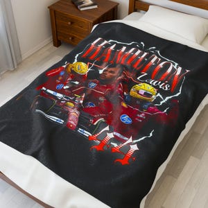 May include: Black blanket featuring a graphic of a Formula 1 race car and driver. The blanket has the name "HAMILTON" in red, stylized text. The driver is wearing a red racing suit and yellow helmet. The blanket is on a bed.