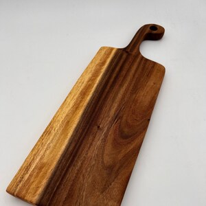 May include: A long, rectangular wooden serving board with a handle. The board is made of dark brown wood with a natural grain pattern. The handle is a simple loop shape.