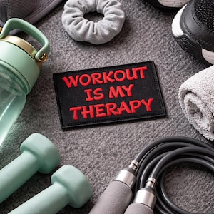 Könnte beinhalten: Eine Auswahl an Fitnessgeräten, darunter Kurzhanteln, eine Wasserflasche und ein Springseil, auf einer grauen Oberfläche angeordnet. Ein schwarzer Aufnäher mit den roten Worten "WORKOUT IS MY THERAPY" ist ebenfalls vorhanden. Schwarz-weiße Turnschuhe sind sichtbar.