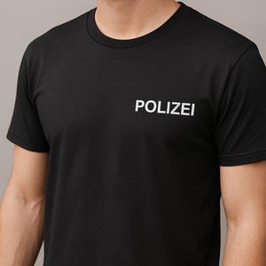 Könnte beinhalten: Ein schwarzes kurzärmeliges T-Shirt mit Rundhalsausschnitt und dem Wort POLIZEI in weißen Großbuchstaben auf der linken Brustseite. Das Kleidungsstück besteht aus einfarbigem schwarzem Stoff mit einer Standardpassform und einem schlichten Rundhalsdesign.
