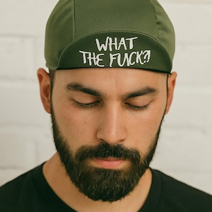 Könnte beinhalten: Olivgrüne Fahrradkappe mit dem weißen Text "WHAT THE FUCK?!", getragen von einer Person mit dunklem Bart und schwarzem T-Shirt. Der Hintergrund ist eine weiße Ziegelmauer.