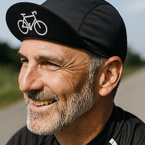 Könnte beinhalten: Eine schwarze Radsportmütze mit einer weißen Strichzeichnung eines Fahrrads auf dem vorderen Schirm. Die Mütze besteht aus einem strukturierten Stoff und hat eine abgerundete Kronenform, die eng am Kopf anliegt und als Kopfbedeckung dient.