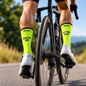 Könnte beinhalten: Ein Paar neongelbe Radsocken mit einem schwarzen Bergsymbol am oberen Rand und dem Text "CATCH ME" auf der linken Socke sowie "IF YOU CAN" auf der rechten Socke. Die Socken werden mit weißen Radschuhen auf einem schwarzen Fahrrad getragen.