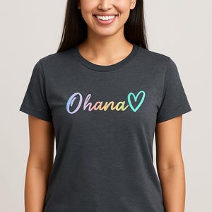 Könnte beinhalten: Dunkelgraues T-Shirt mit dem Wort "Ohana" in farbenfroher, verlaufender Schrift und einem türkisfarbenen Herzsymbol. Das Shirt hat einen Rundhalsausschnitt und scheint aus weichem Material zu sein.