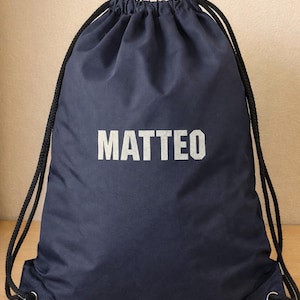 Könnte beinhalten: Marineblauer Turnbeutel mit dem Namen "MATTEO" in weißen Blockbuchstaben. Die Tasche hat schwarze Kordelzüge und Metallösen. Der Beutel liegt auf einer Holzoberfläche.