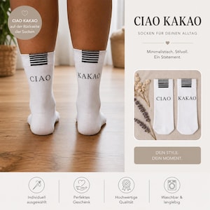 Könnte beinhalten: Ein Paar weiße Socken mit schwarzen horizontalen Streifen am oberen Rand und dem Schriftzug CIAO auf der linken sowie KAKAO auf der rechten Socke. Die Socken werden an Unterschenkeln und flach liegend in einem kleinen Bildausschnitt gezeigt.