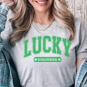 May include: A gray t-shirt with the word "LUCKY" in large green letters, and the word "ENGINEER" below it, with a shamrock symbol. The shirt is worn with a denim jacket and dark jeans.