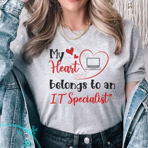 May include: A light gray t-shirt with the text "My Heart belongs to an IT Specialist" in red and black. The design includes a heart with a laptop inside. The shirt is a casual style.