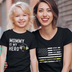 May include: Two black t-shirts with a white and yellow design. The smaller shirt says "Mommy is my hero" with a 911 symbol and a stethoscope. The larger shirt has an American flag with a yellow stripe across the bottom that says "911 Dispatcher" and the words "Mommy Protector Hero" above the stripe.