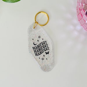 May include: A clear, glittery keychain with a gold ring. The keychain is shaped like a motel key and has the word "MANIFEST" repeated three times in black text. There is a small crescent moon and a butterfly graphic on the keychain.