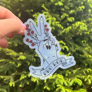 K&ouml;nnte beinhalten: Ein blauer Aufkleber mit einer Hand, die ein Peace-Zeichen macht und einen Zweig mit roten Blumen h&auml;lt. Der Aufkleber hat ein Banner mit der Aufschrift "PRO ROE" in schwarzen Buchstaben.