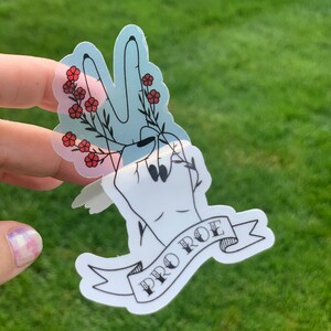 May include: A sticker with a black outline of a hand making a peace sign with red flowers around the fingers. The sticker has a banner with the text 'PRO ROE' in black lettering.