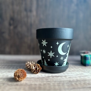 May include: Black ceramic planter with a white crescent moon and snowflake design. The planter has a matching saucer.