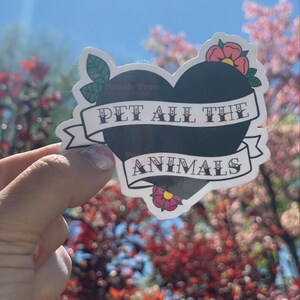 May include: A black heart-shaped sticker with the text "PET ALL THE ANIMALS" in white lettering. The sticker has a white banner with a red flower on each side.