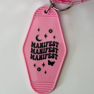 May include: Pink plastic hotel key chain with the word "MANIFEST" repeated three times in black lettering. The keychain also features a crescent moon, a butterfly, and stars.
