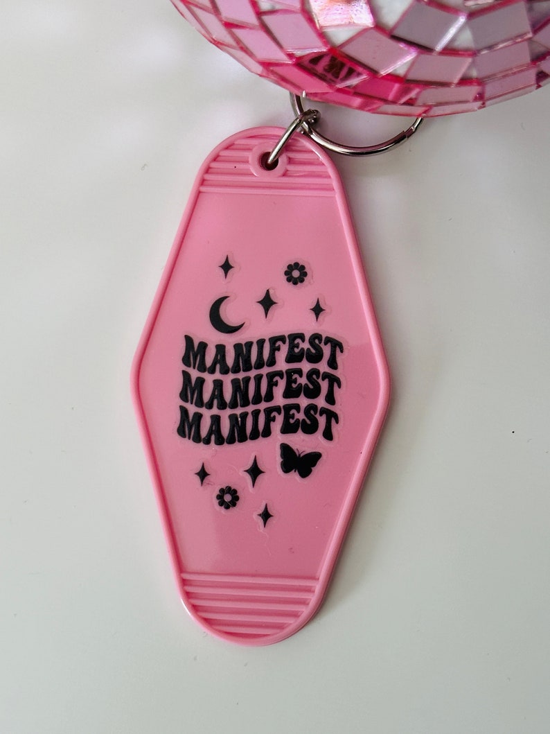 May include: Pink plastic hotel key chain with the word "MANIFEST" repeated three times in black lettering. The keychain also features a crescent moon, a butterfly, and stars.