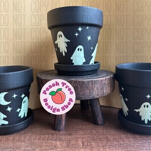 May include: Three black terracotta pots with white ghost and star designs. The pots are on a wooden stand with a pink sticker that says "Peach Tree Design Shop".