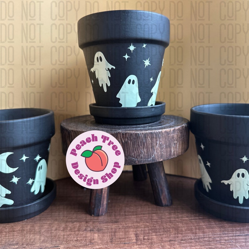 May include: Three black terracotta pots with white ghost and star designs. The pots are on a wooden stand with a pink sticker that says "Peach Tree Design Shop".