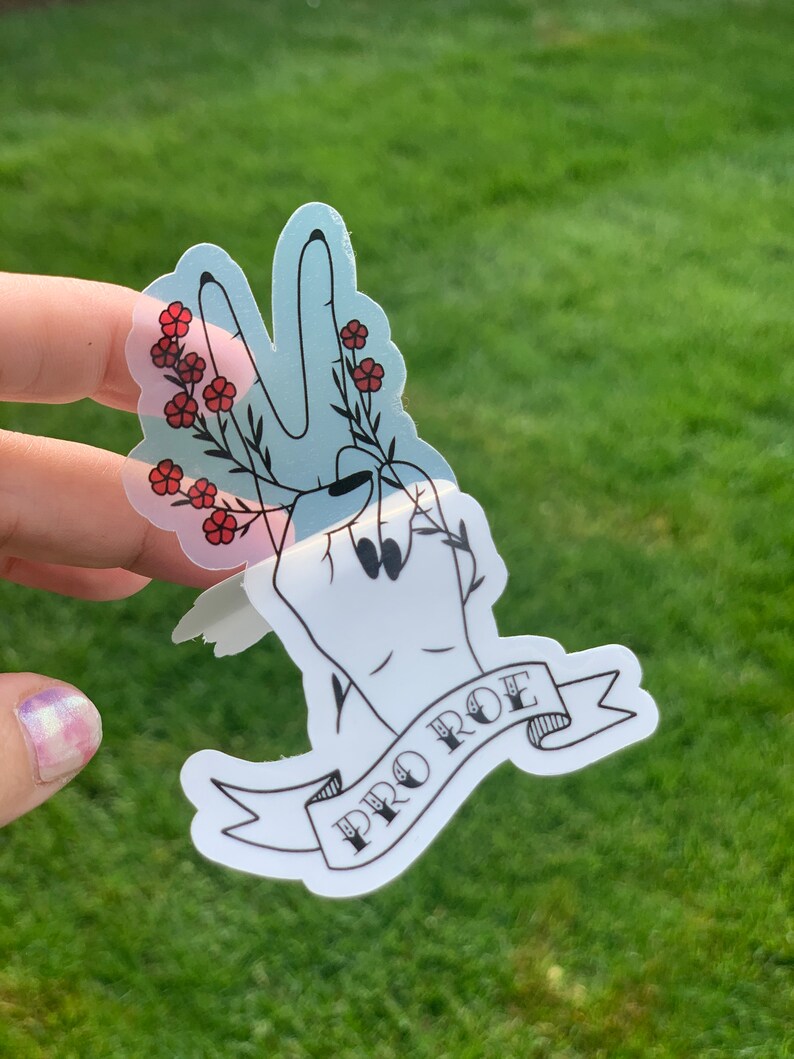 May include: A sticker with a black outline of a hand making a peace sign with red flowers around the fingers. The sticker has a banner with the text 'PRO ROE' in black lettering.