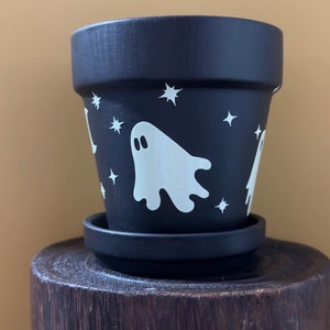 May include: Black terracotta planter with a white ghost and stars design. The planter has a matching saucer.