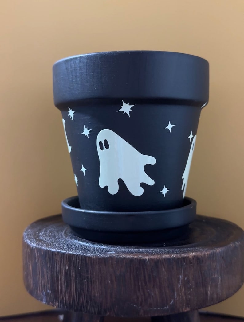May include: Black terracotta planter with a white ghost and stars design. The planter has a matching saucer.