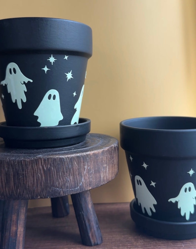 May include: Two black terracotta flower pots with white ghost and star designs. The pots are on a wooden stool and a wooden surface.